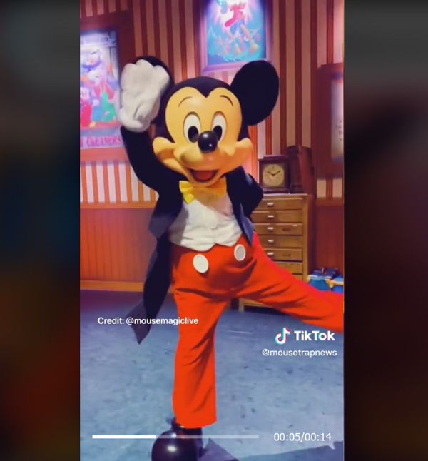 A Mickey Mouse mascot wearing a yellow bowtie, white shirt, black jacket, red pants with two white buttons, and black shoes is waving with its right leg kicked out. 