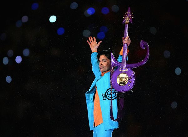 A Black man in a turquoise suit and orange shirt holds his hands up, one holding a bright purple guitar.