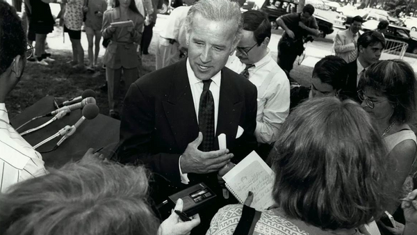 A white man wearing a black suit talks to people who are holding recorders and microphones. There are other people standing behind the man.