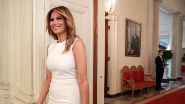 A white woman wearing a white dress smiles as she stands in a hallway.
