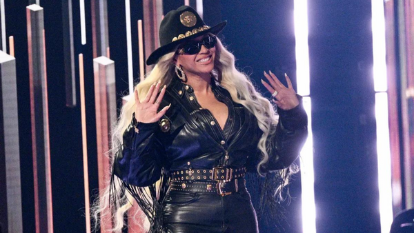 A Black woman wearing a black cowboy hat, black sunglasses, and black leather jacket and pants waves.