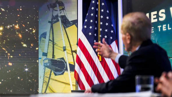 A white man points towards an image of a telescope next to an American flag.