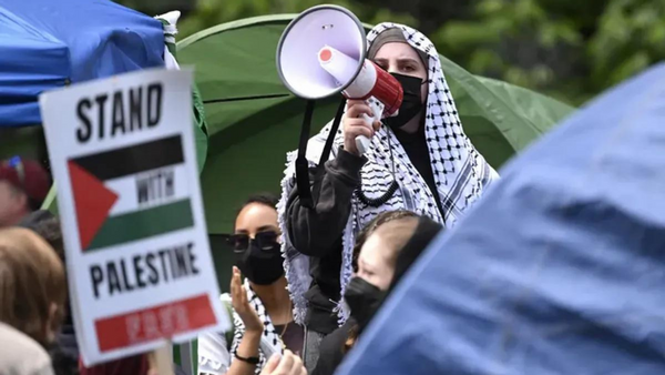 A person wearing a mask and a keffiyeh holds a bullhorn up to their mouth. People stand around them. A sign that says STAND WITH PALESTINE can be seen on the left side of the photo.