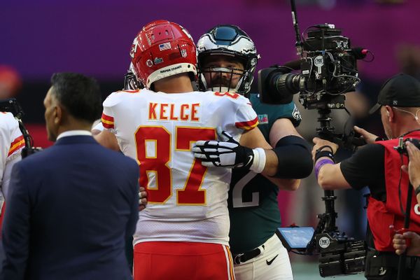 Two white men hug surrounded by people. One is wearing a red jersey that says "Kelce 87," while the other is wearing a green helmet. A reporter is interviewing a person to the left side of the photo, while a cameraperson is on the right side of the two.
